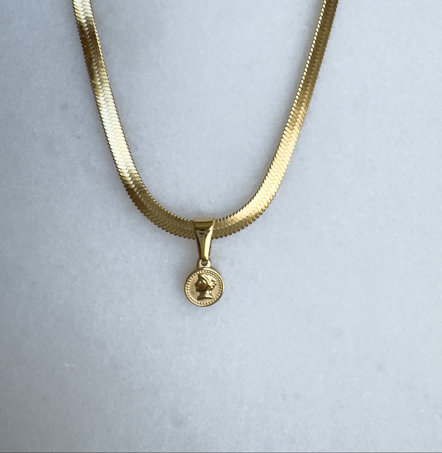 Gold herringbone necklace with a round pendant on a light gray background