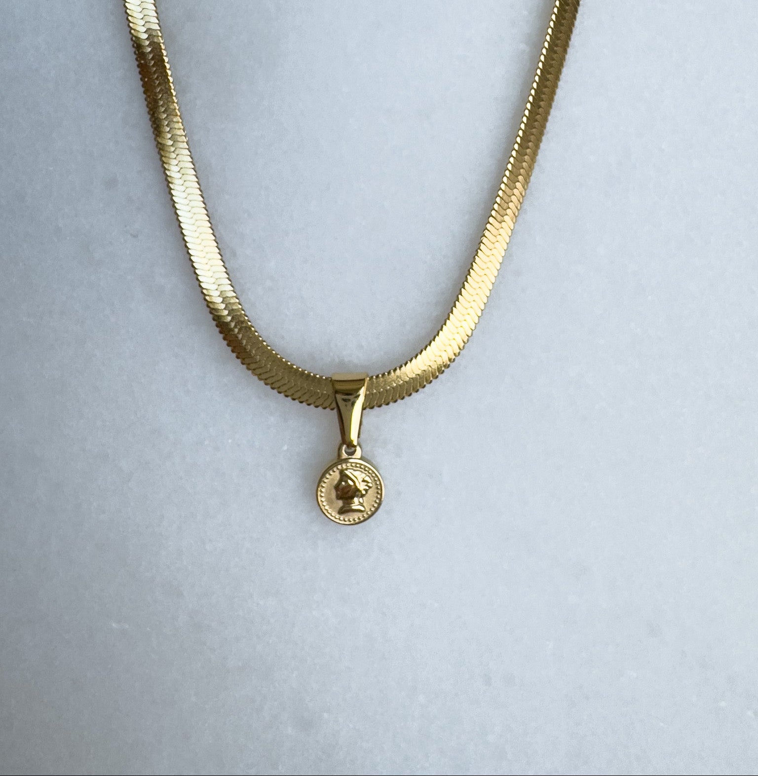 Gold herringbone necklace with a round pendant on a light gray background